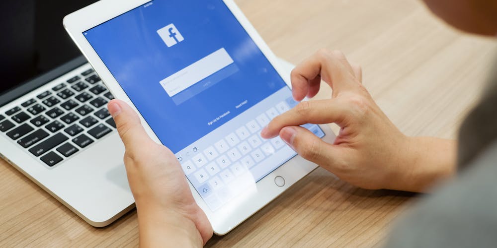 Person sitter foran laptop med ipad i hendene og logger inn på facebook