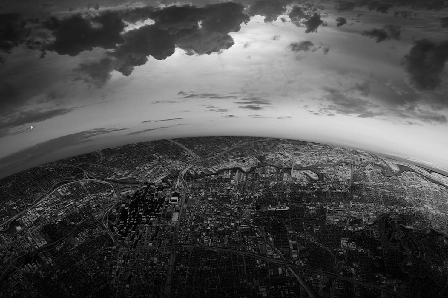 Sort/hvitt-bilde tatt høyt ovenfra, hvor jorden og en by kurver rundt på sidene og skyene er mørke på himmelen