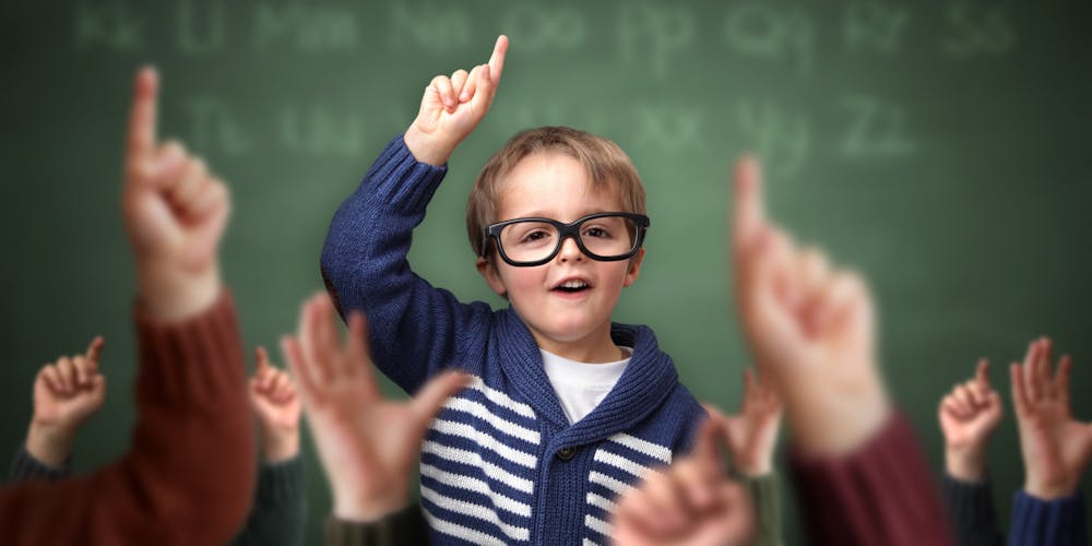 Ung gutt med briller foran tavle rekker opp hånden. Mange andre barnehender er også rukket opp.