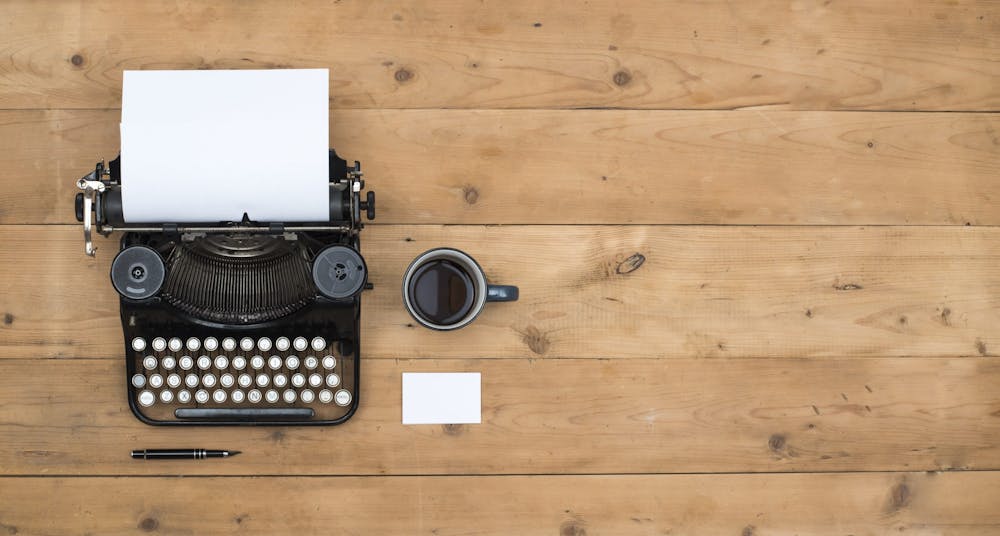 Bilde av en gammel skrivemaskin og en kopp kaffe på et trebord. Illustrerer at arbeid med tekst er viktig for å nå ut til markedet.