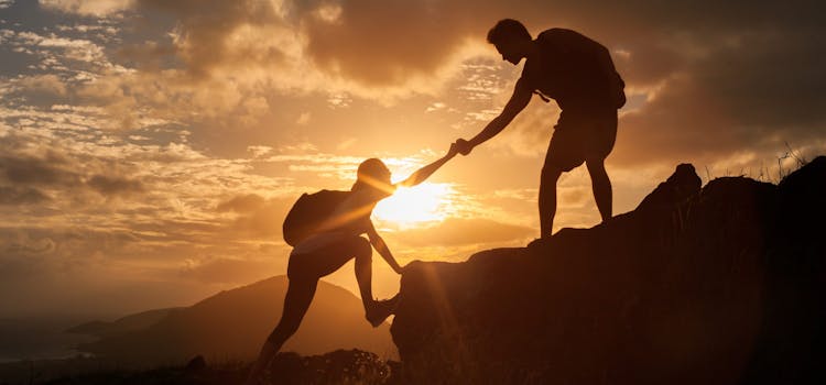 Siluett av to mennesker som hjelper hverandre opp et fjell i solnedgang.